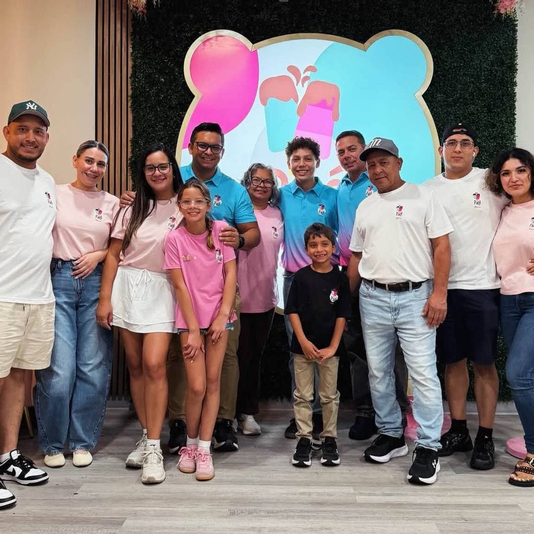 La familia y el equipo de Palé —alrededor de una docena de personas con camisetas de Palé— frente a la pared de flores y el letrero redondo de Palé