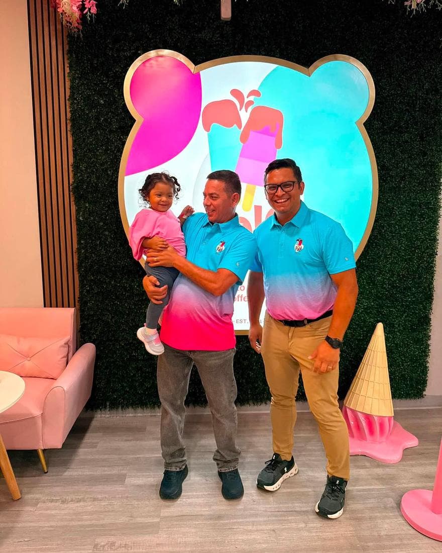 Alejandro and Elder in Palé polo shirts in front of the flower wall, one of them holding a small child