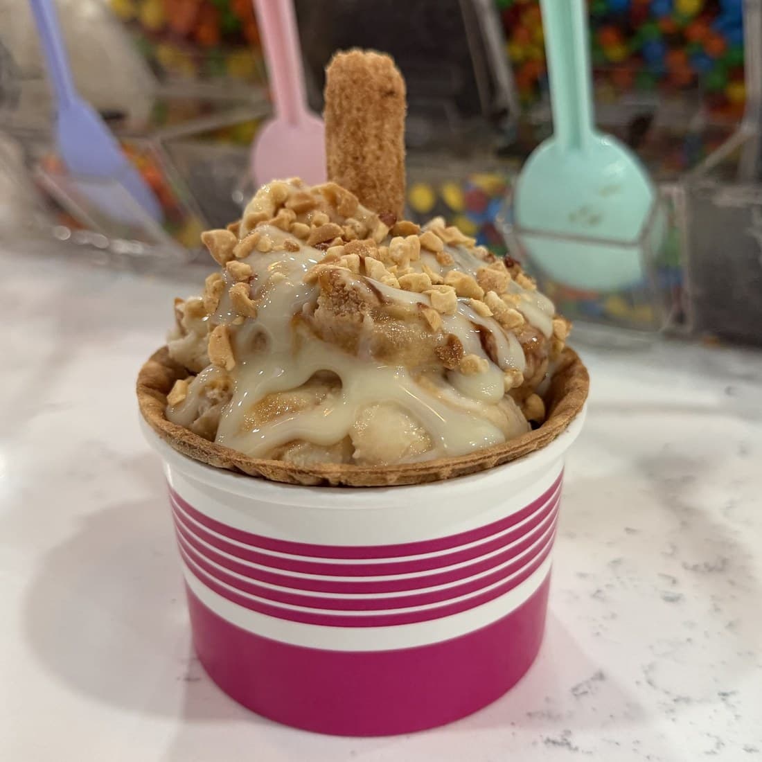 Un vaso de gelato de Palé con caramelo y frutos secos triturados, en un vaso a rayas magenta, junto a la barra de toppings