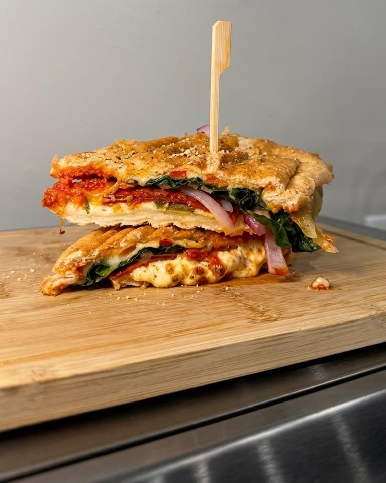 A toasted Palé panini cut in half and stacked, layered with tomato sauce, melted cheese, greens and red onion, a bamboo pick through the top, on a wooden board